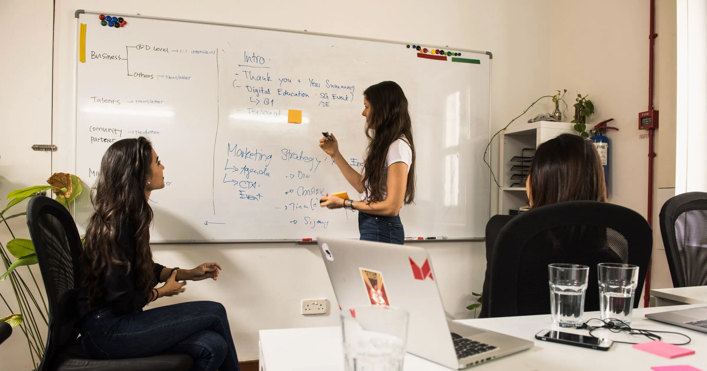 Three colleagues in a meeting room, one writing a marketing strategy on a whiteboard while the others watch
