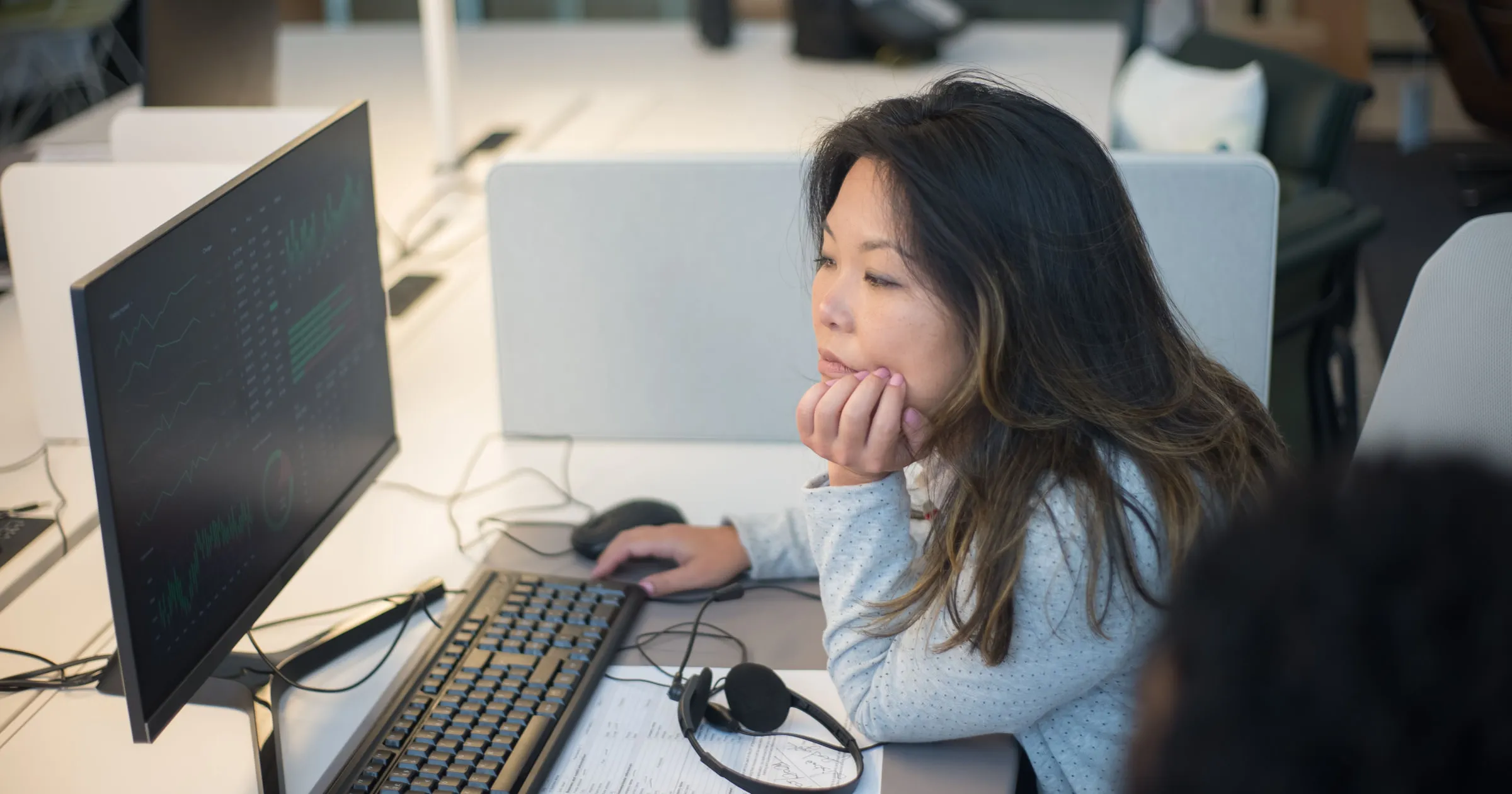 A woman at her desk, chin in her hand, studying charts on her computer monitor